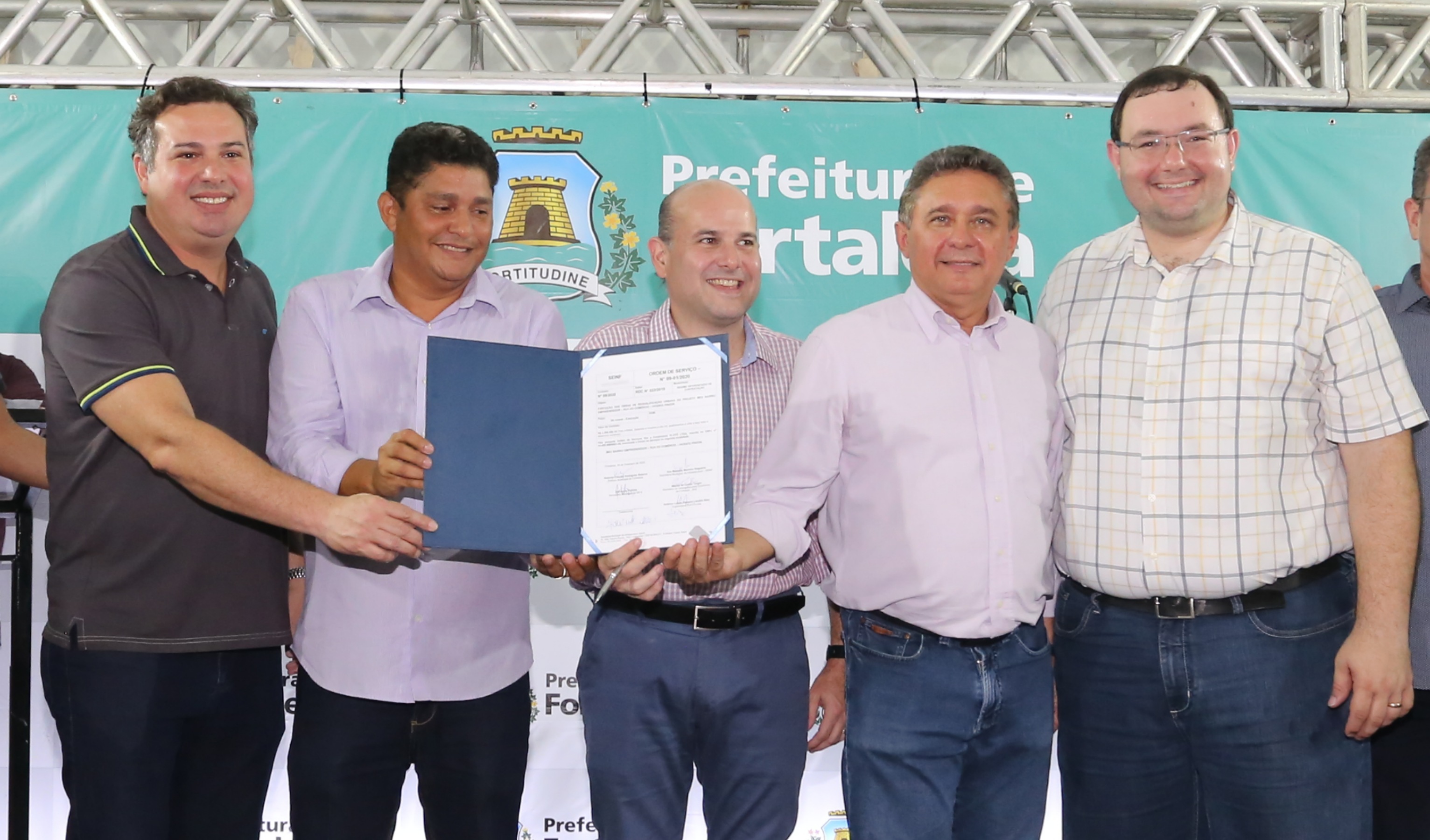 prefeito e senhora em cima de palco segurando documento e sorrindo para a foto